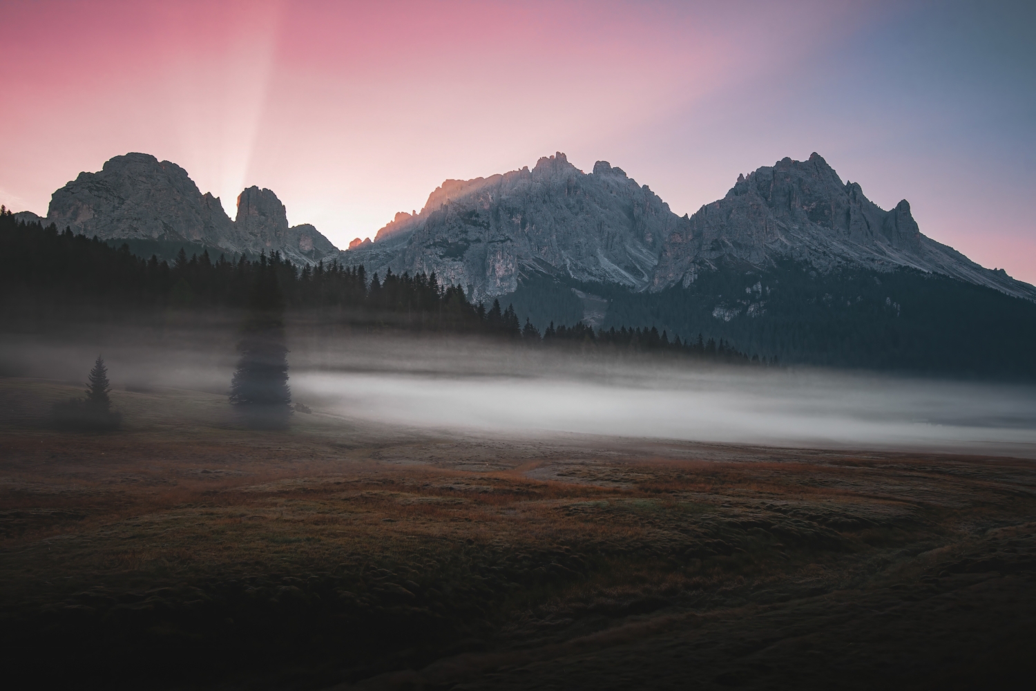 Dolomiten-Landschaft am frühen Morgen, mit ersten Sonnenstrahlen hinter den Bergen, Bodennebel und rot-violettem Himmel
