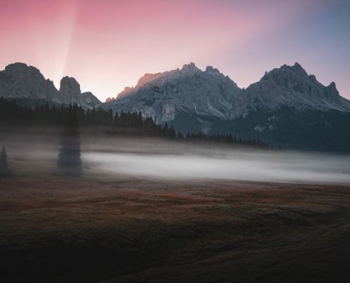 Dolomiten-Landschaft am frühen Morgen, mit ersten Sonnenstrahlen hinter den Bergen, Bodennebel und rot-violettem Himmel