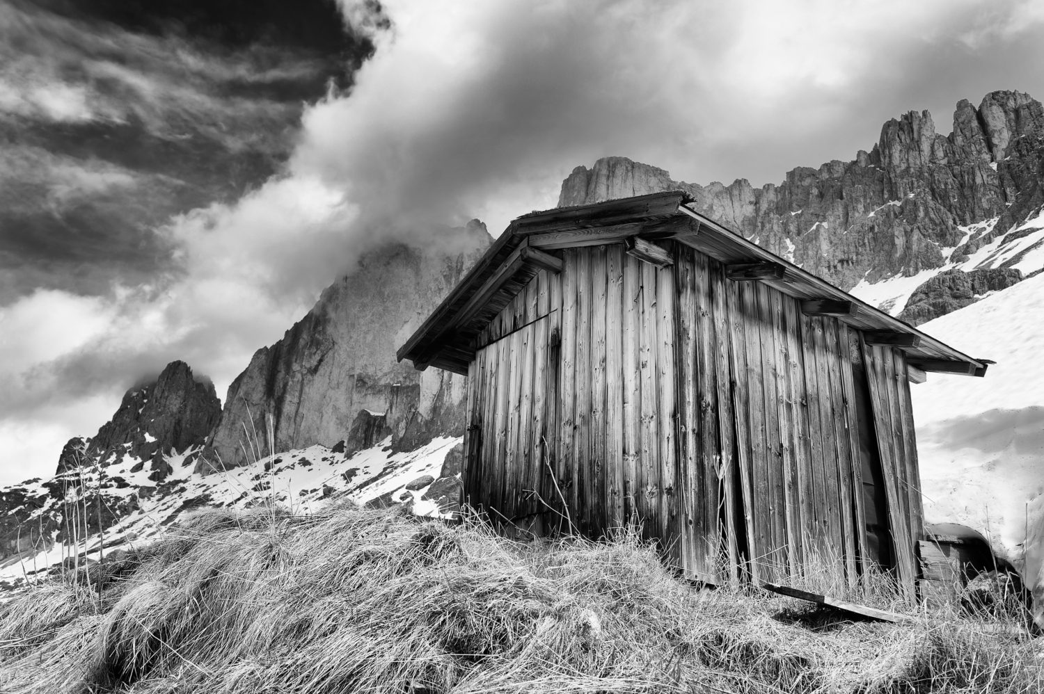 Schwarzweiss-Bild einer stimmungsvollen hochalpinen, teils verschneiten Berglandschaft, aus der Froschperspektive mit Holzhütte