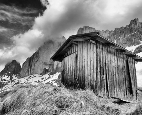Schwarzweiss-Bild einer stimmungsvollen hochalpinen, teils verschneiten Berglandschaft, aus der Froschperspektive mit Holzhütte