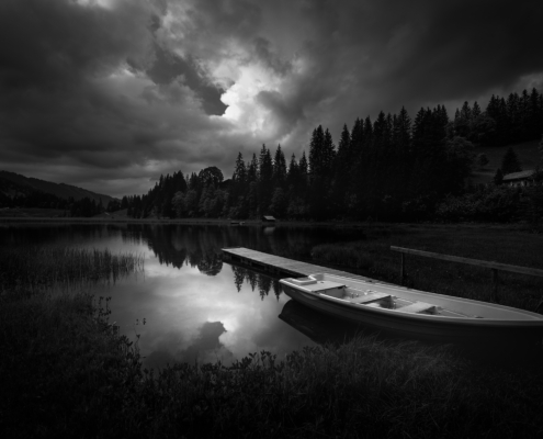 Von Wolken düster-dramatisch beleuchtete, ruhige, Landschaftsszene mit Boot in schwarzweiss am Lauenensee (CH)